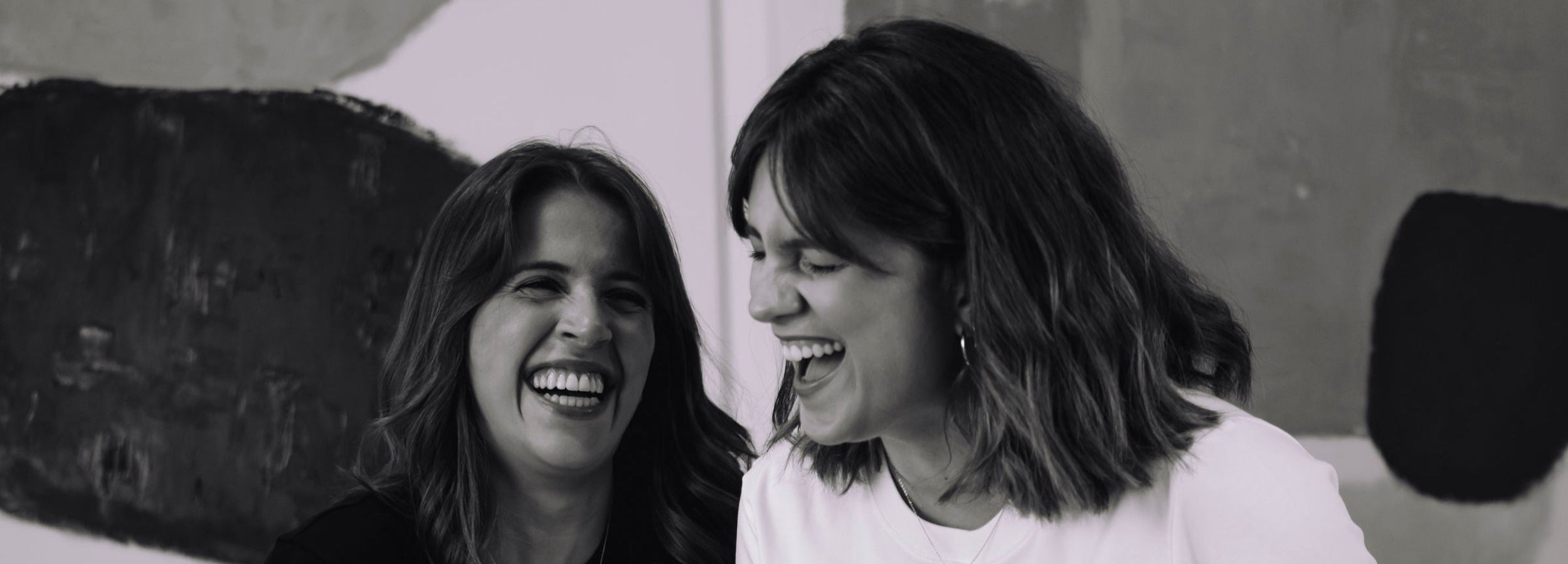 Two women laughing together at a laptop in a room with abstract art on the walls.