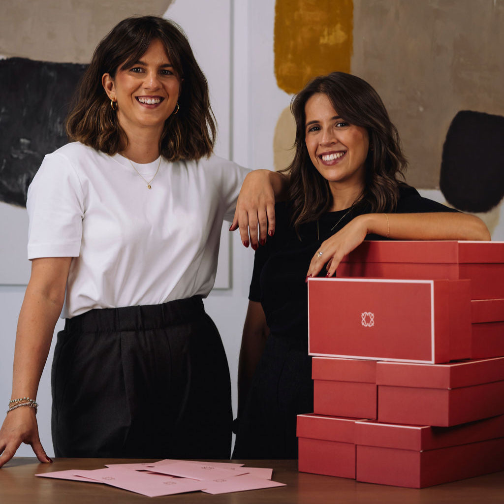 Two women standing behind a table with red boxes and pink cards in an indoor setting.