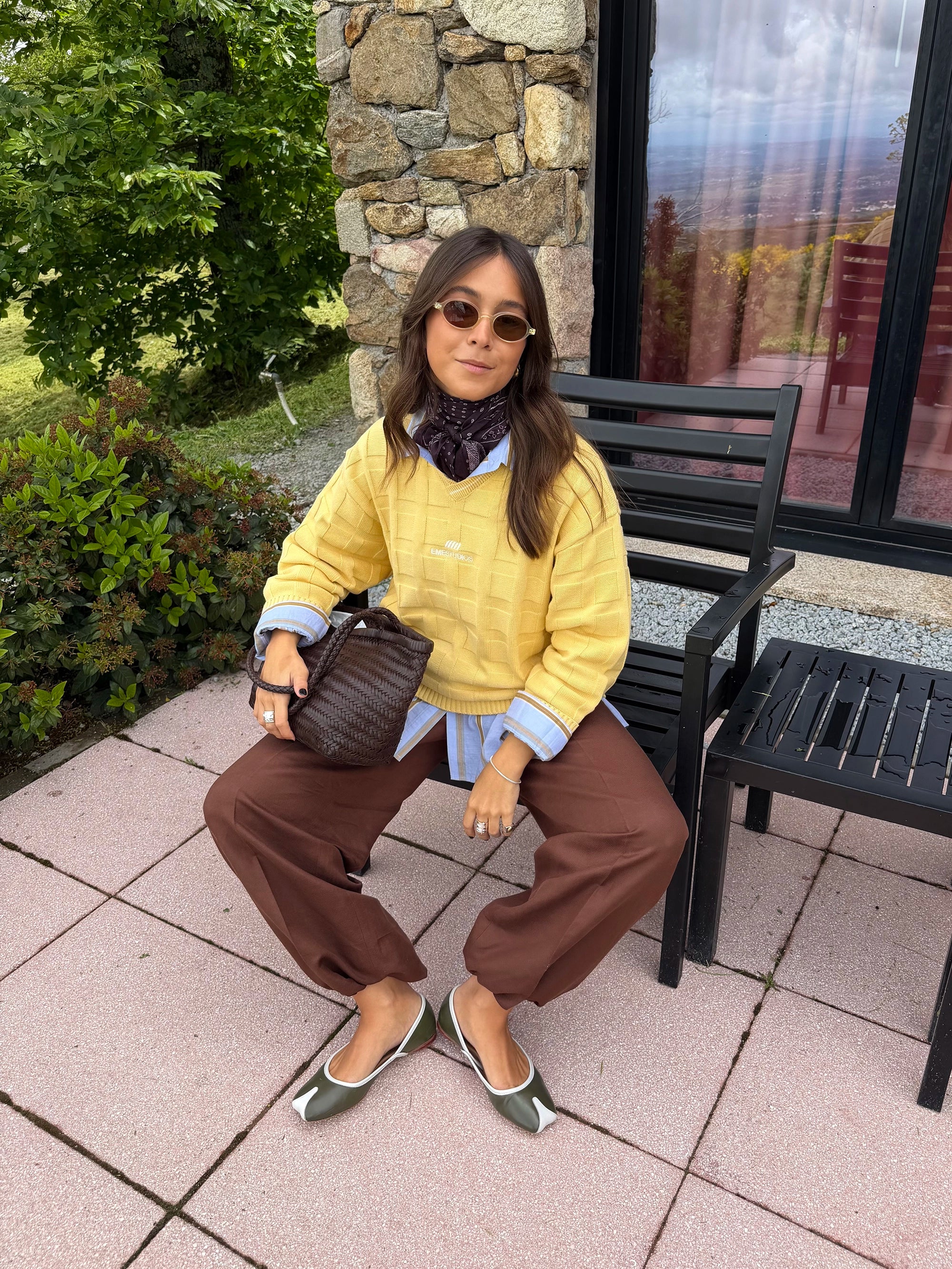 Woman in yellow sweater and brown pants sitting outdoors on a patio.