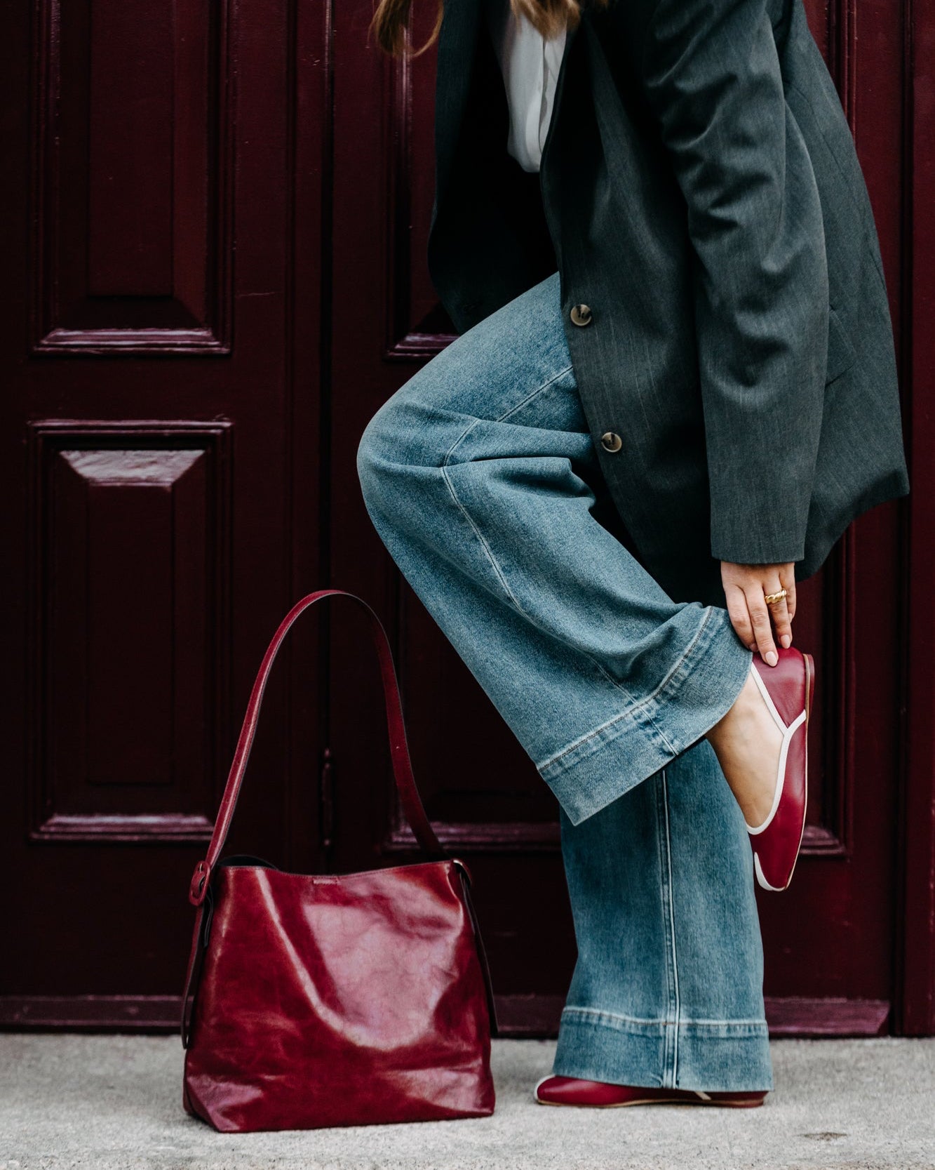 Person wearing a dark coat, blue jeans, and red shoes with a maroon handbag against a red door.