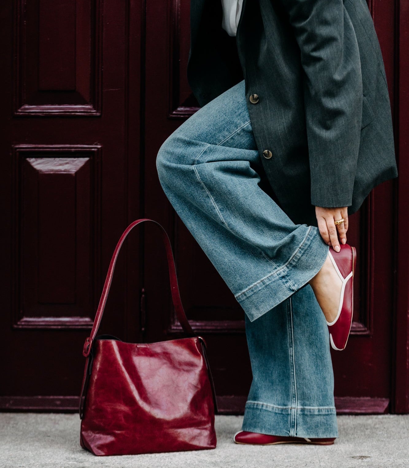 Person wearing a dark coat, blue jeans, and red shoes with a maroon handbag against a red door.