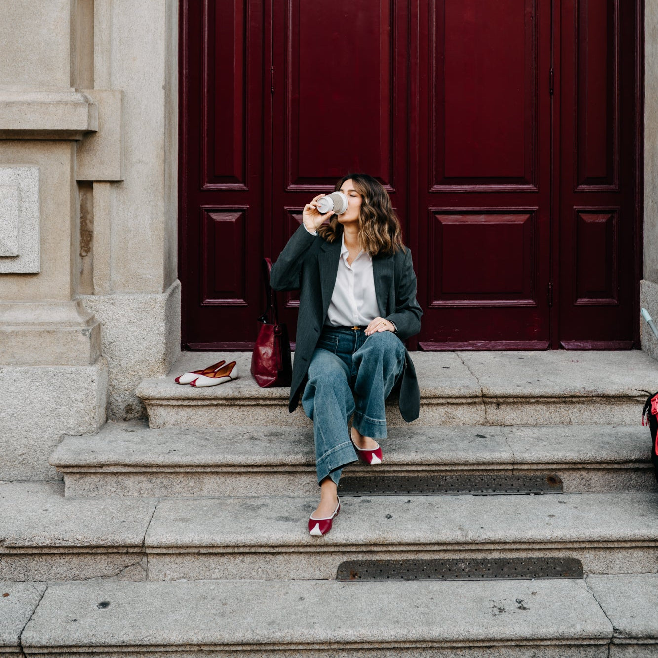 Woman sitting on steps in front of a large red door, drinking from a cup.