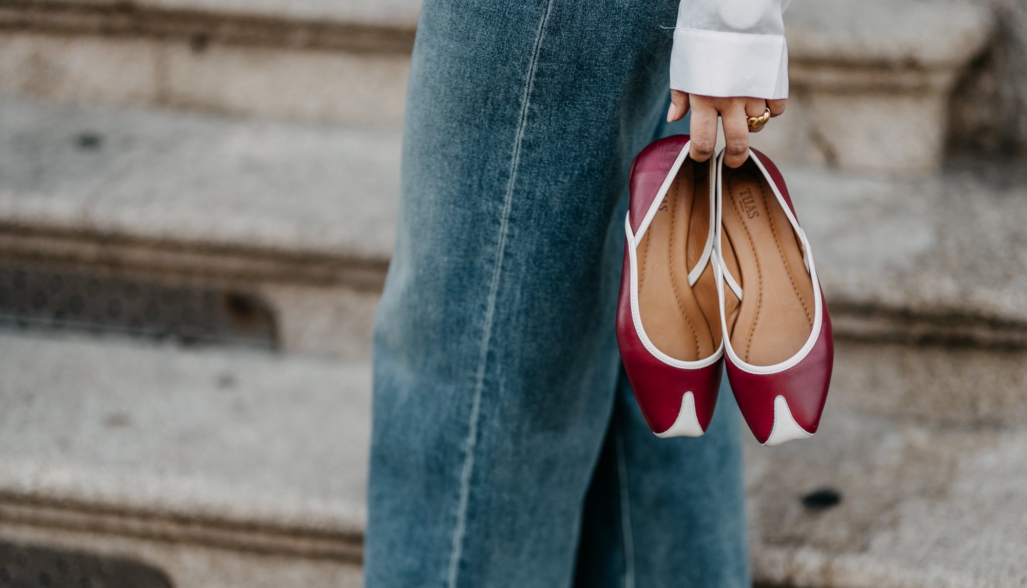 Red and white flat TUAS shoes held by a person wearing blue jeans on stone steps.
