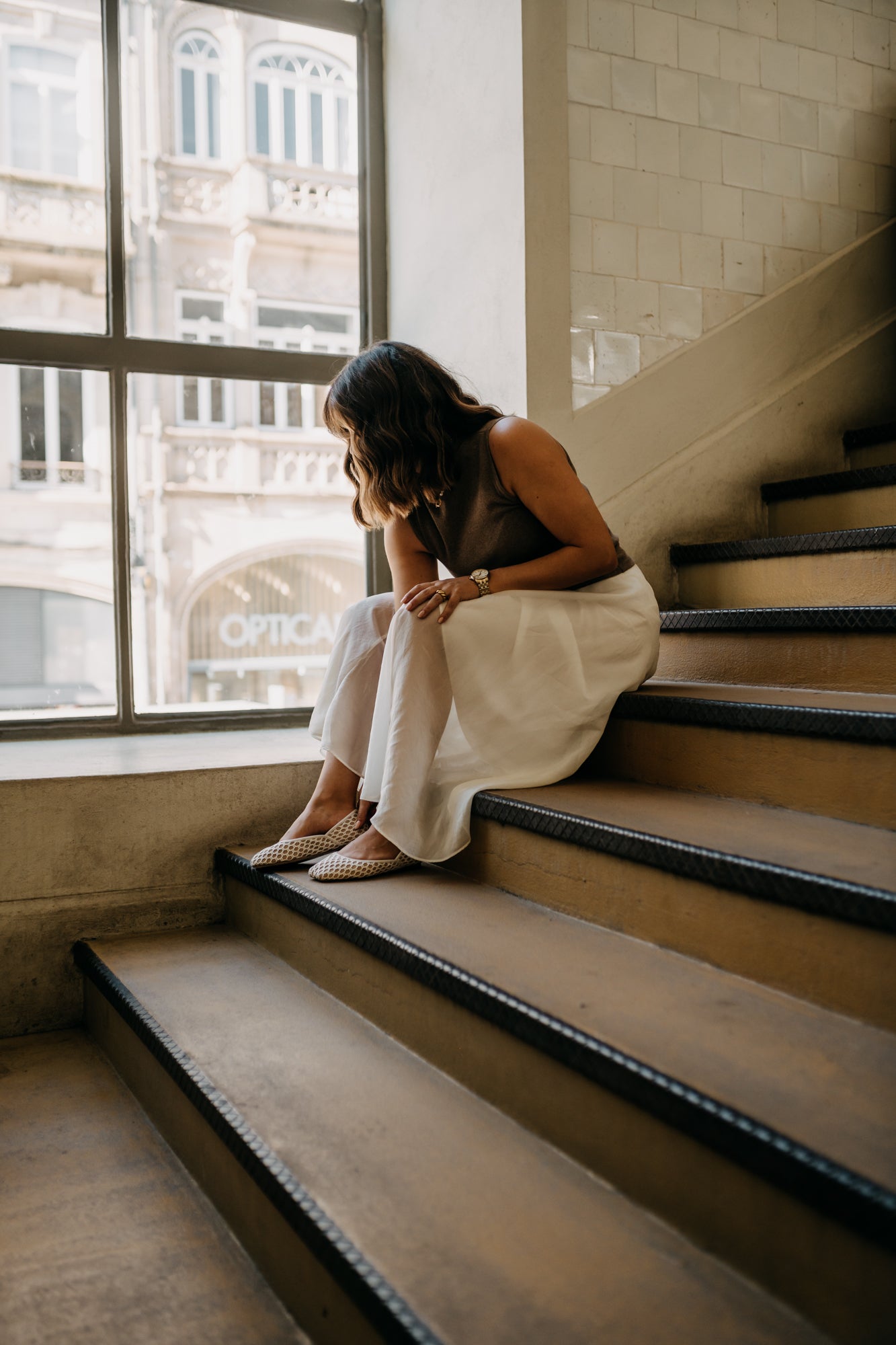 Woman sitting on a staircase in an urban setting with large windows.