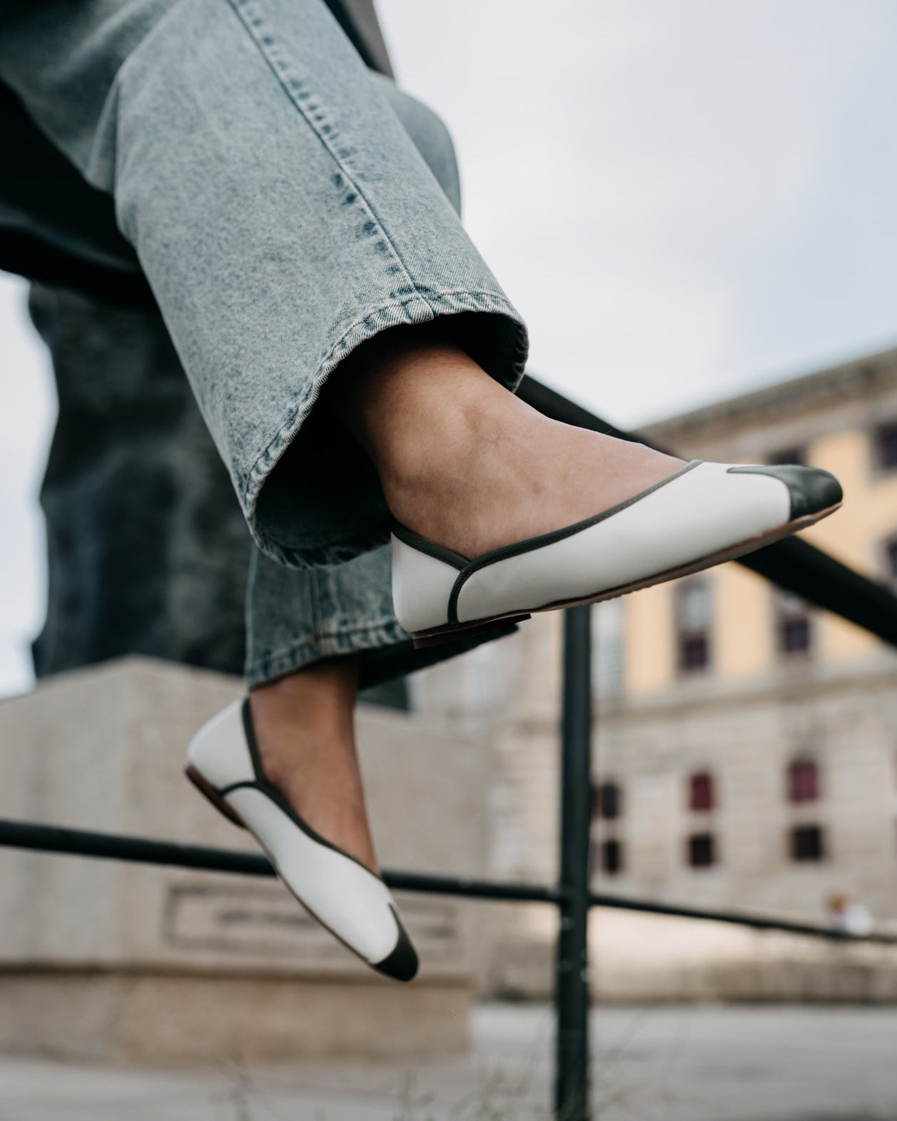 Person wearing black and white flat shoes with jeans, standing on a ledge.
