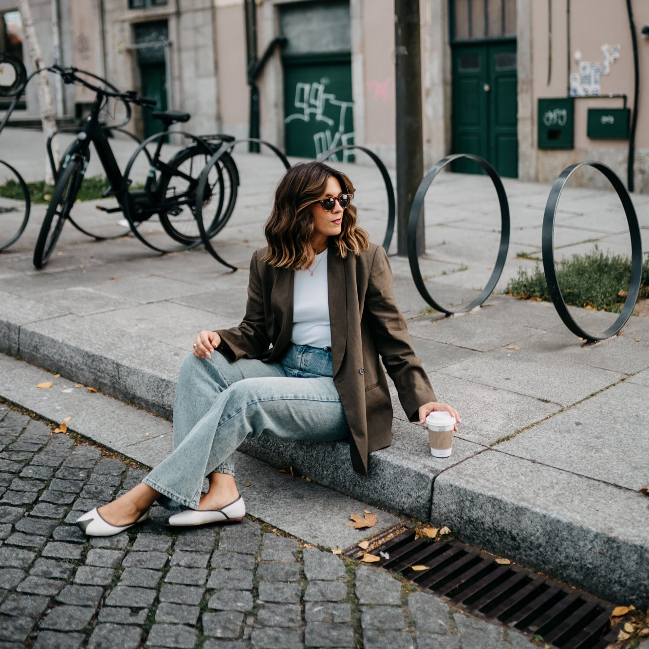 Woman sitting on a city street curb with a coffee cup, surrounded by bicycles and urban architecture.