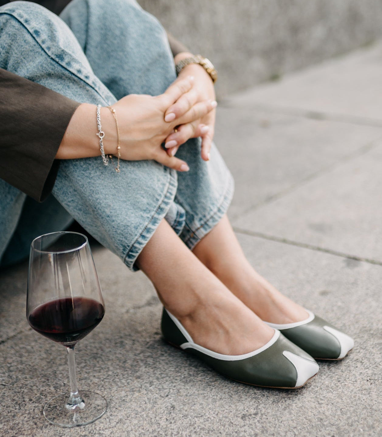 Person sitting on a concrete surface wearing green and white flats with a glass of red wine.