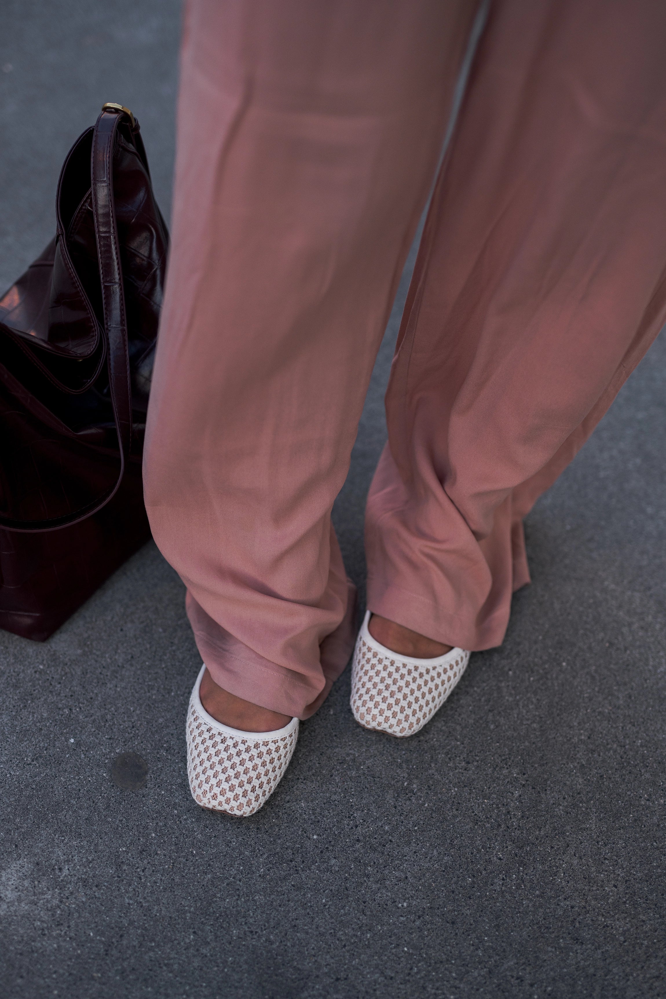 A person wearing dusty rose wide-leg trousers and white open-toe mesh flat shoes, standing on a gray pavement next to a dark brown leather handbag.