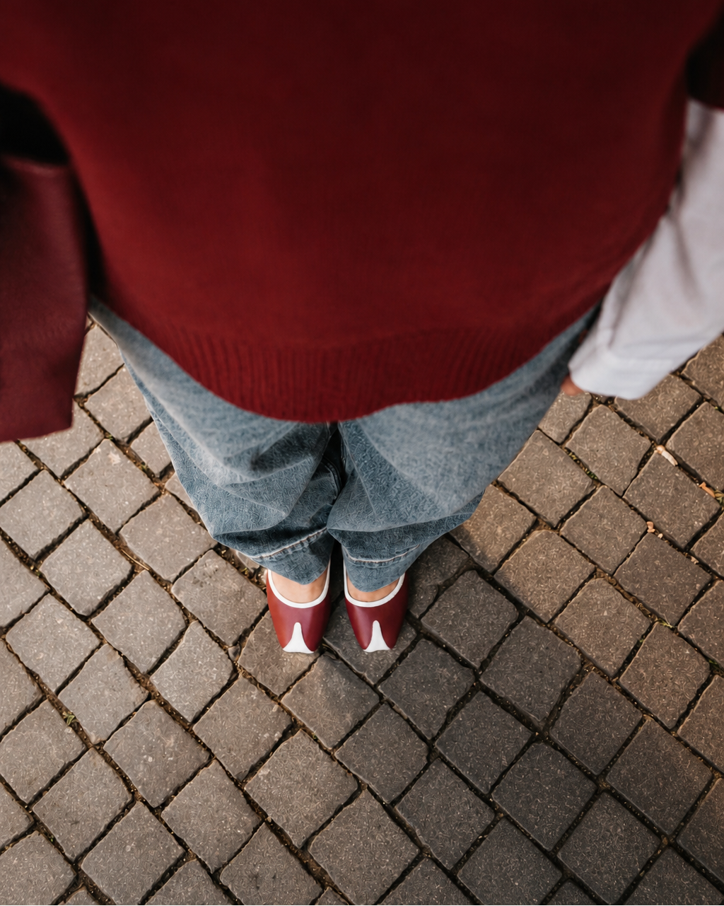 Person wearing a red sweater and blue jeans standing on a brick pavement.