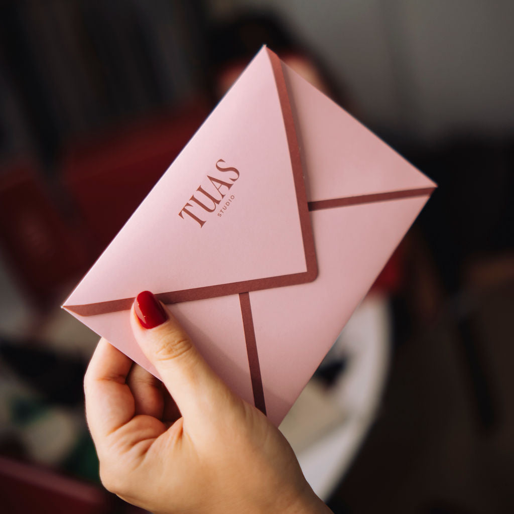 Hand holding a pink envelope with 'TULAS' branding against a blurred background