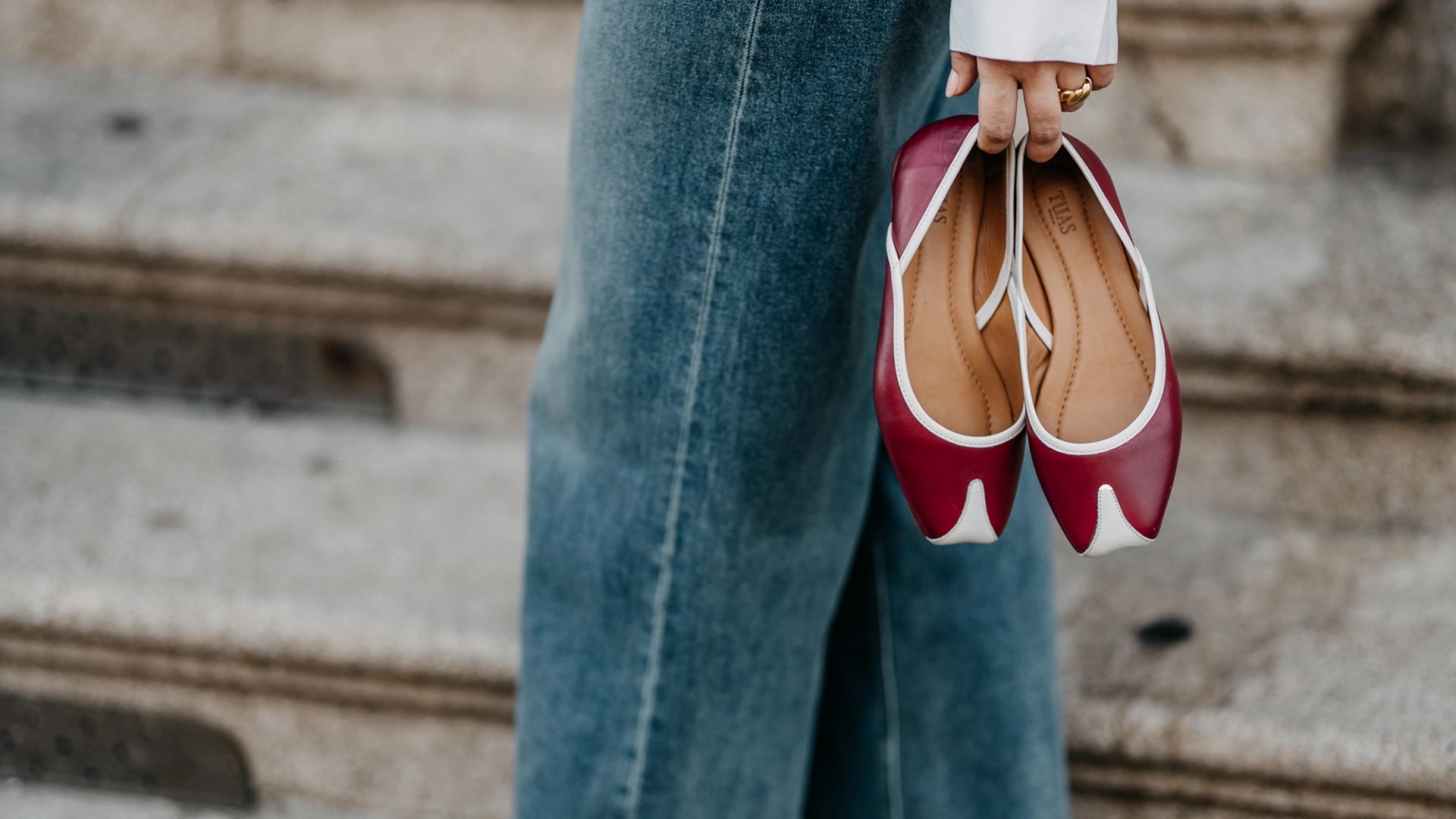 TUAS Julia winery red ballet flats with white soles held by a person wearing blue jeans on stone steps.