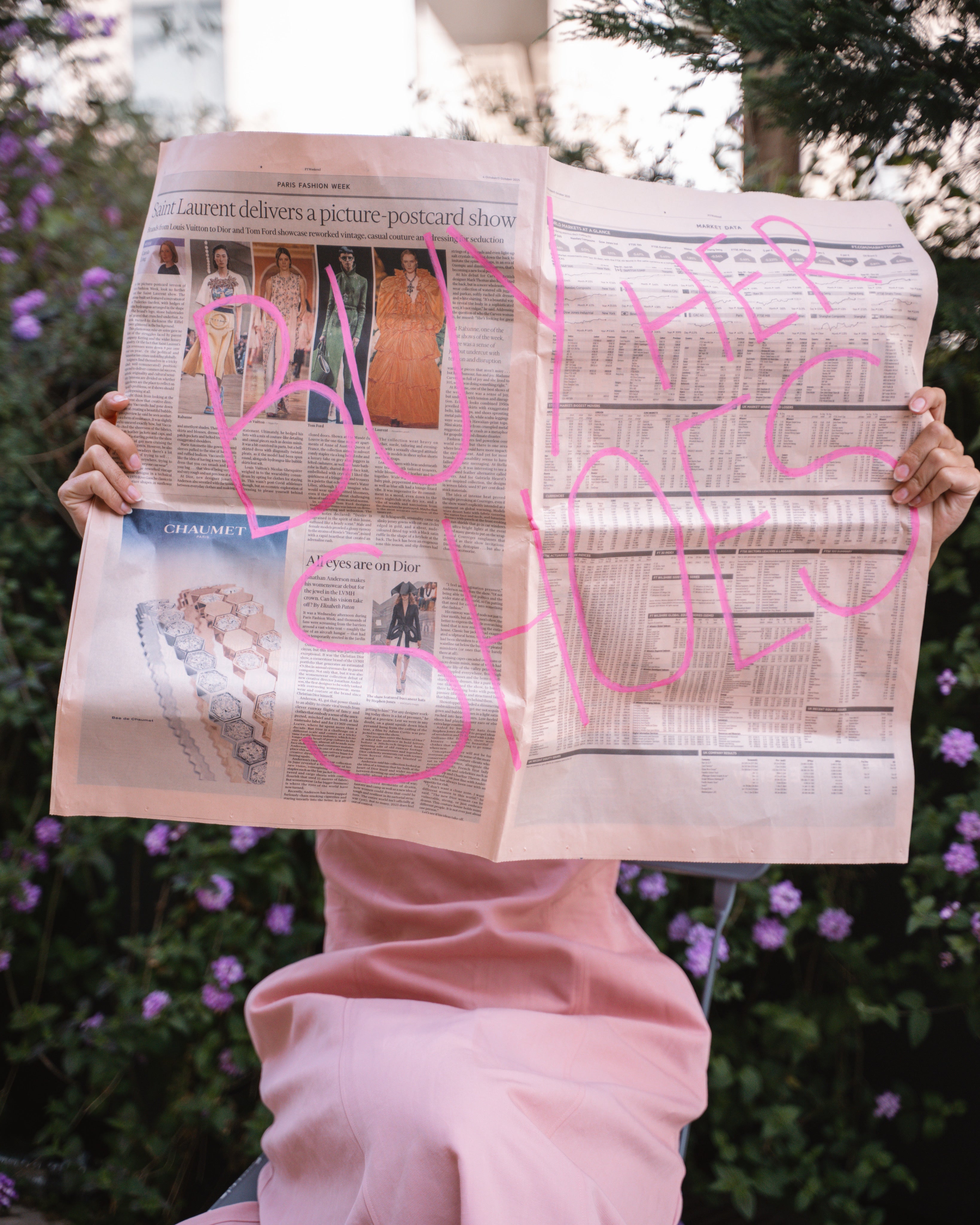 Person holding a newspaper with 'BUY HER SHOES' written on it, outdoors.