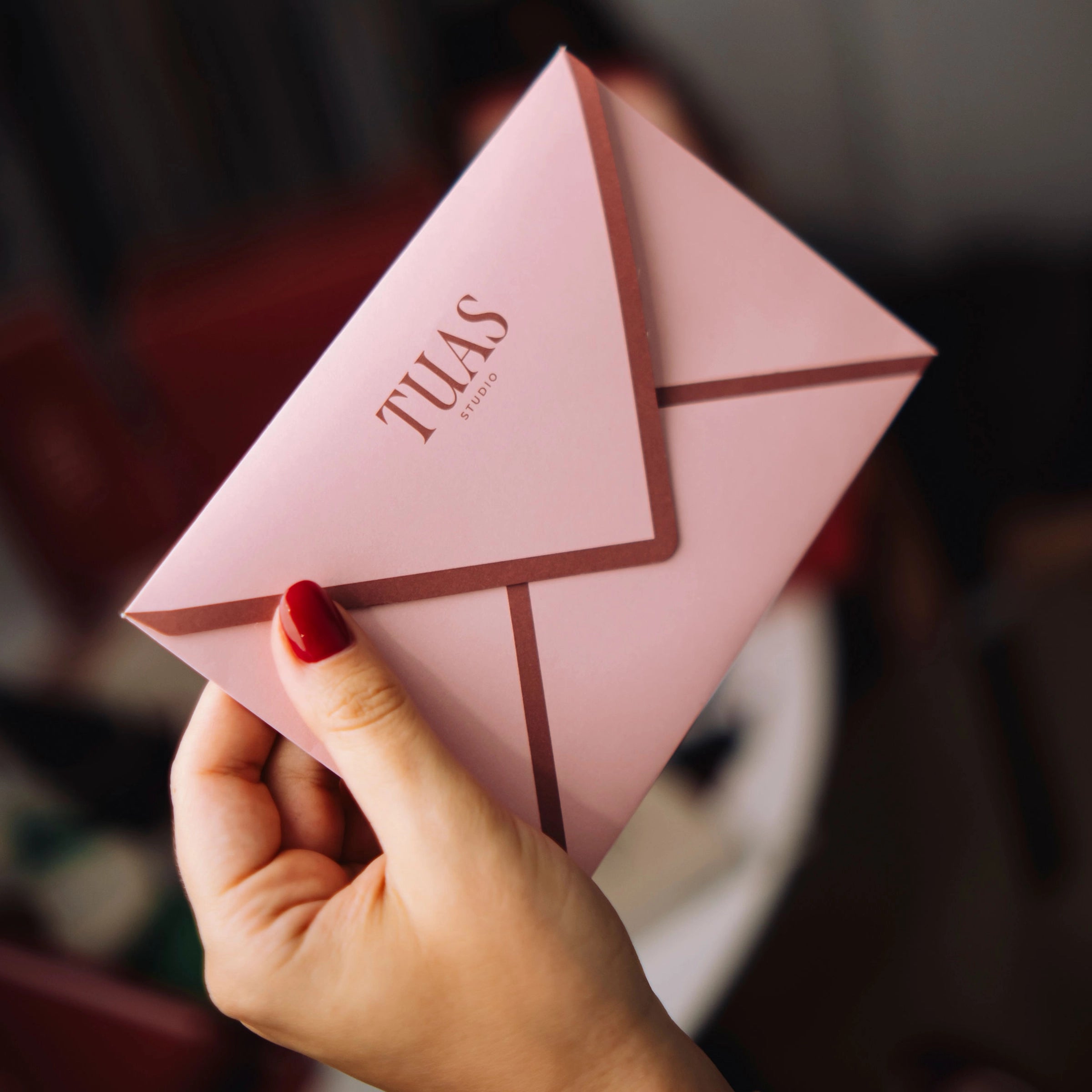 Hand holding a pink envelope with 'TUAS' branding against a blurred background