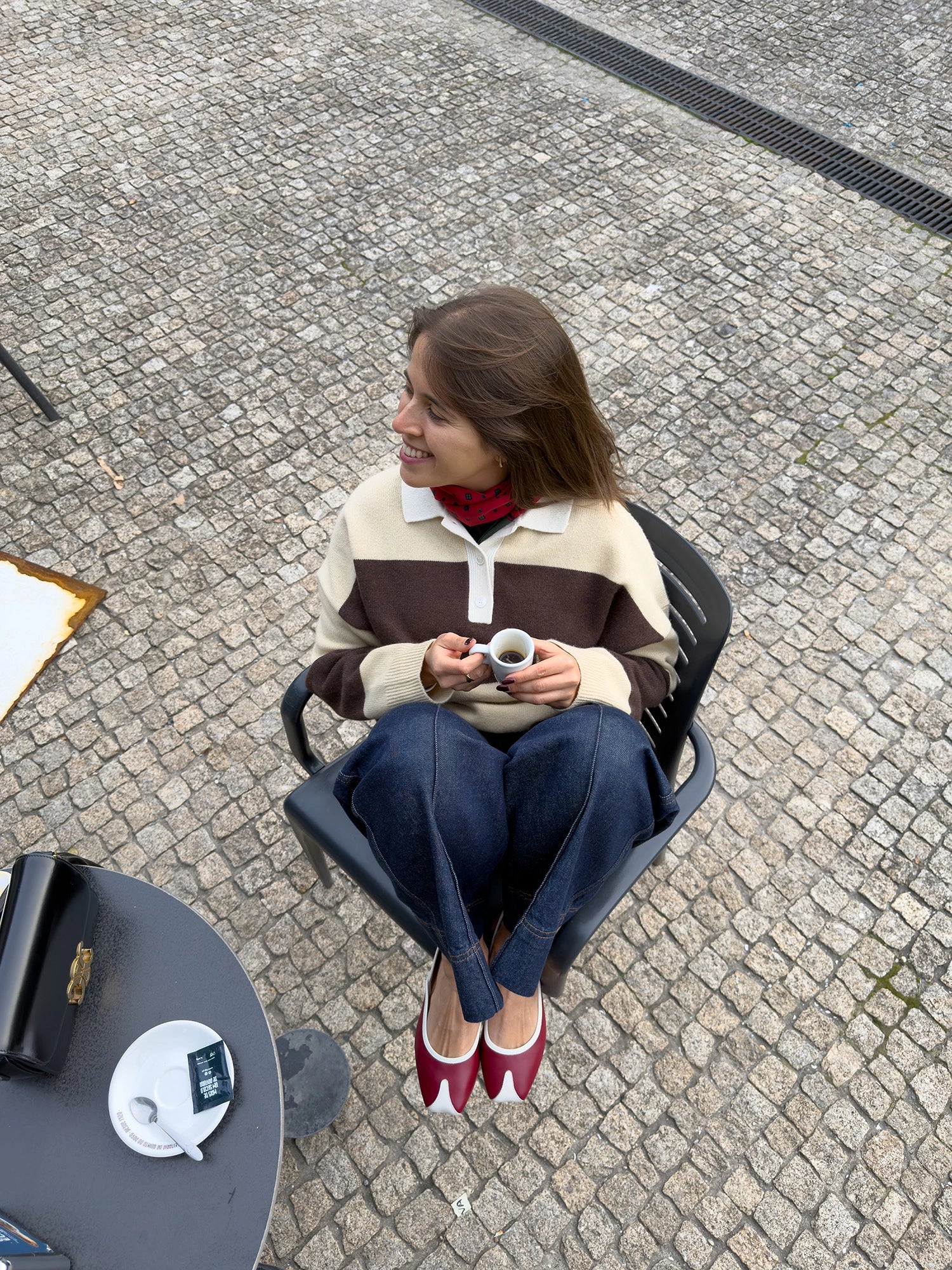 Woman sitting on a chair holding a cup outdoors on a cobblestone street.
