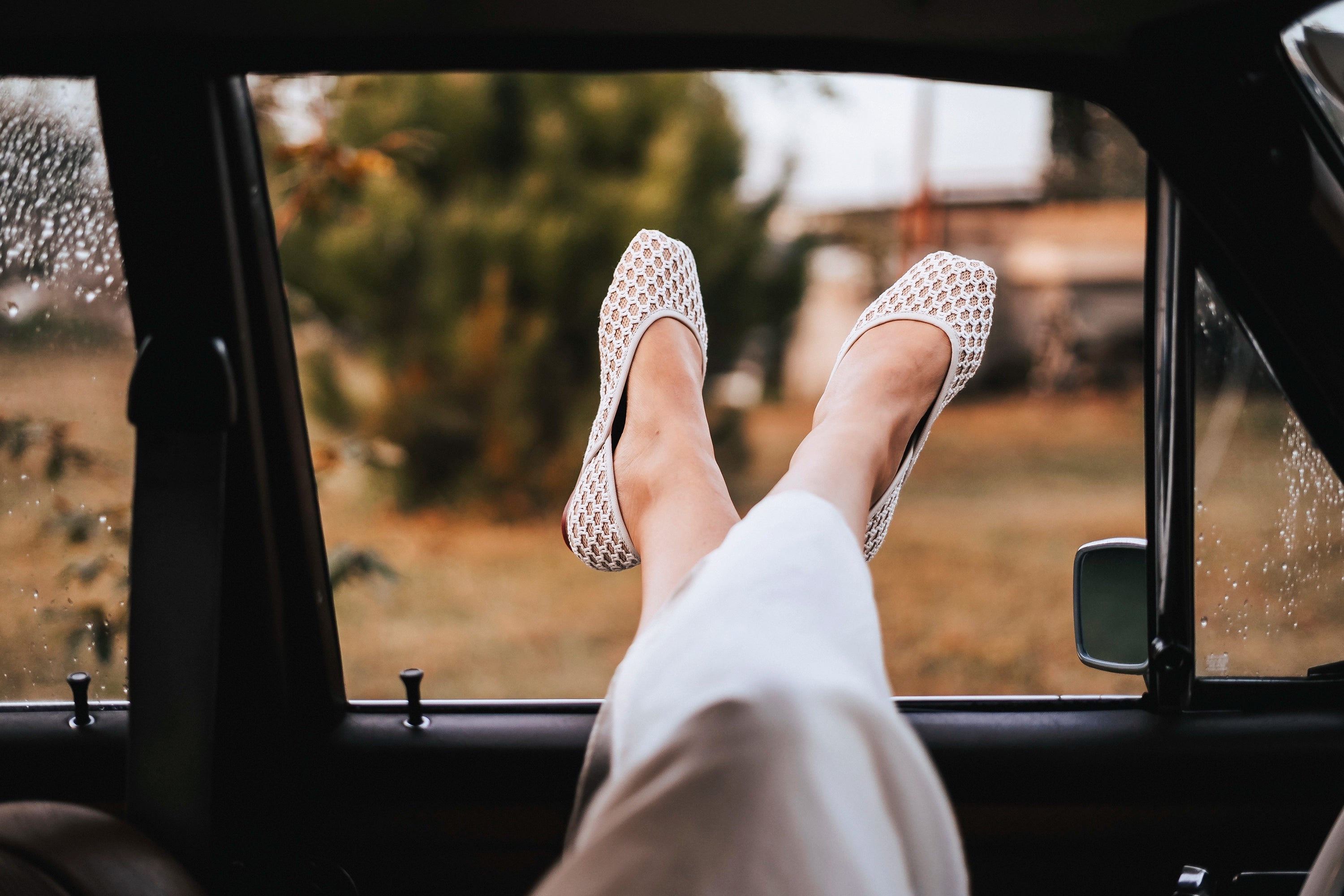 Feet wearing white shoes with a patterned sole, relaxing inside a vehicle with a blurred outdoor scene.