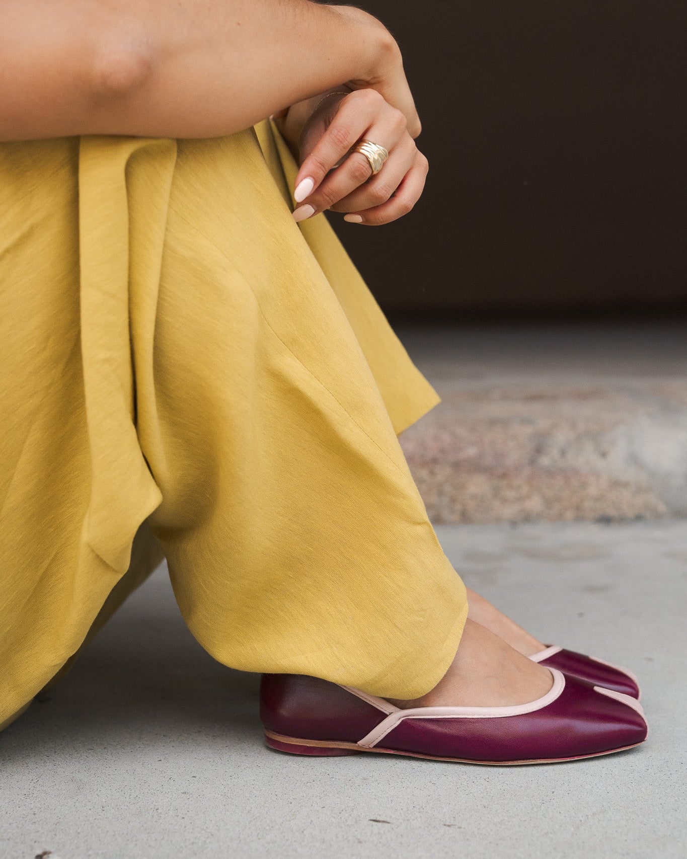 A close-up lifestyle shot of a person sitting on a concrete floor, wearing wide-leg mustard yellow trousers and deep burgundy ballet flats with thin white piping.