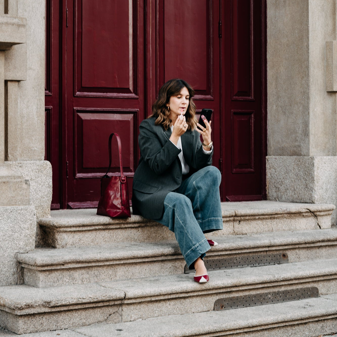 Woman sitting on steps in front of a large red door, using a phone.