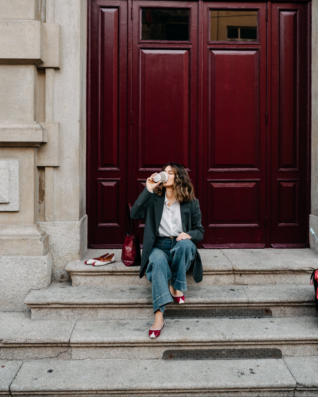 Woman sitting on steps in front of a large red door, drinking from a cup.