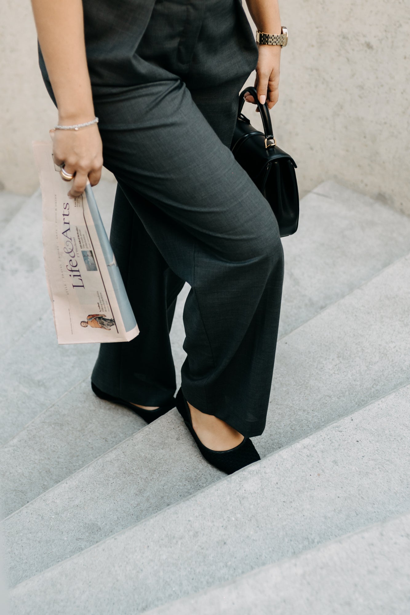 Person in black outfit holding a newspaper and a handbag on a staircase.