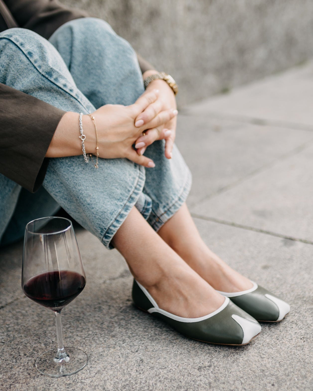 Person sitting on a concrete surface wearing green and white flats with a glass of red wine.
