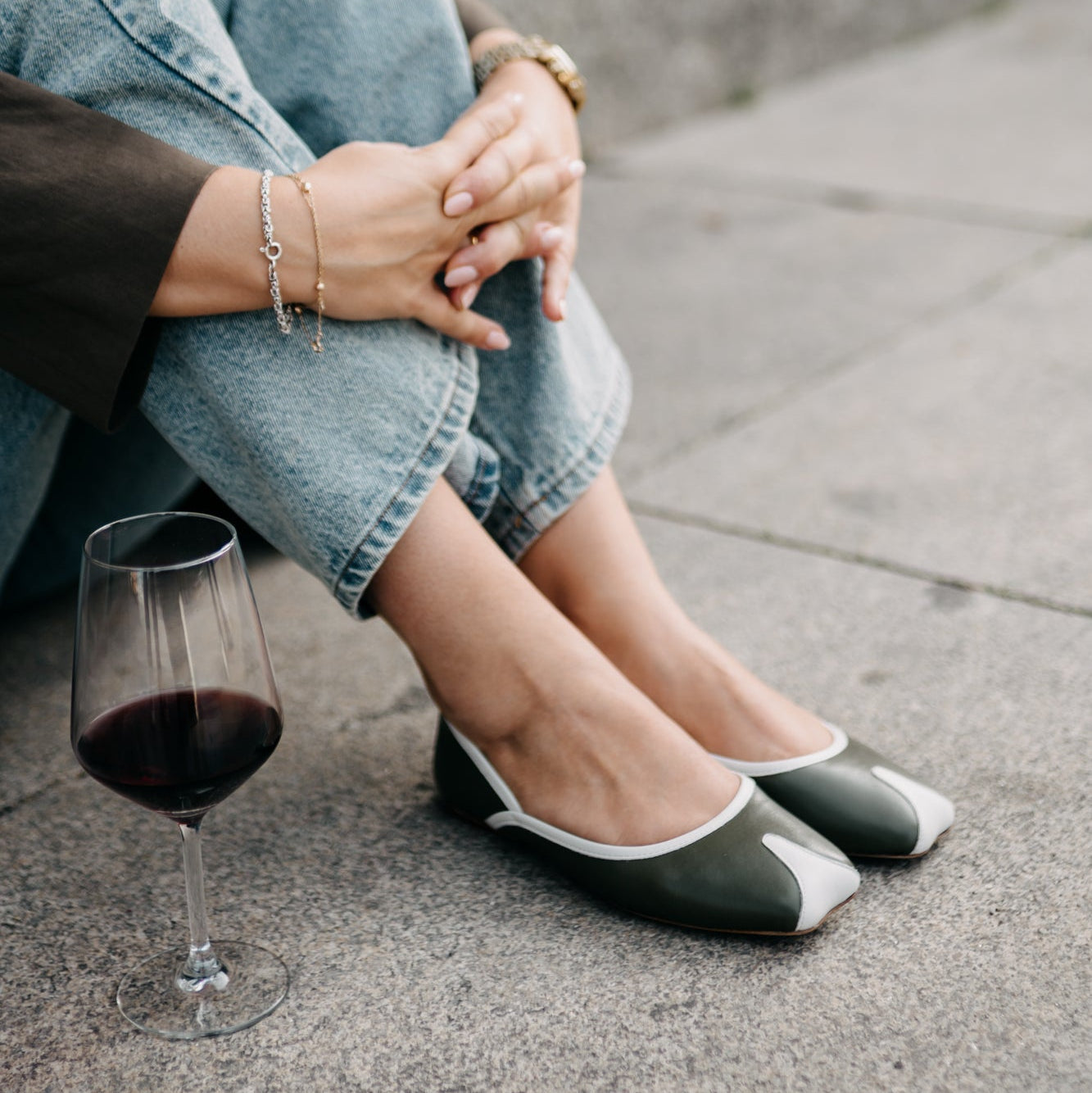 Person sitting on a concrete surface wearing jeans and black flats, with a glass of red wine.